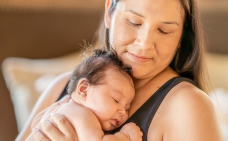 A smiling mother looks down holds her sleeping baby to her chest