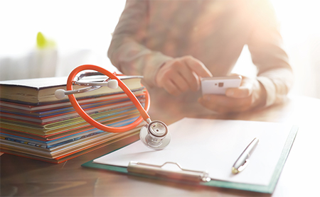 Midsection shot of a doctor sitting at her desk using her phone, in front of her are files, a notepad, and a stethoscope