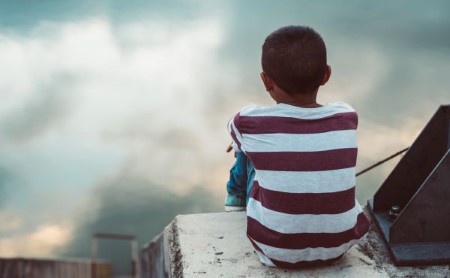 Sad young boy sits on a bridge overlooking the water with his back to the viewer