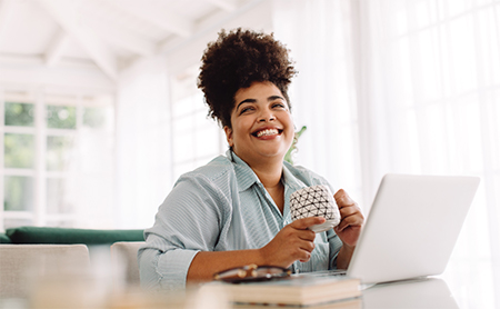 Smiling woman seated at her kitchen island, drinking coffee, working on her laptop