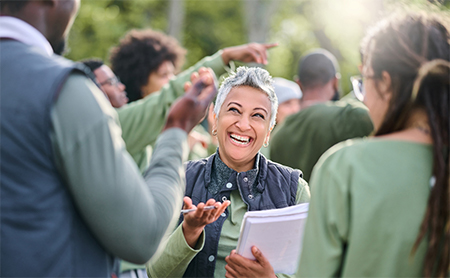 A woman holding a stack of papers talks cheerfully with the people around her